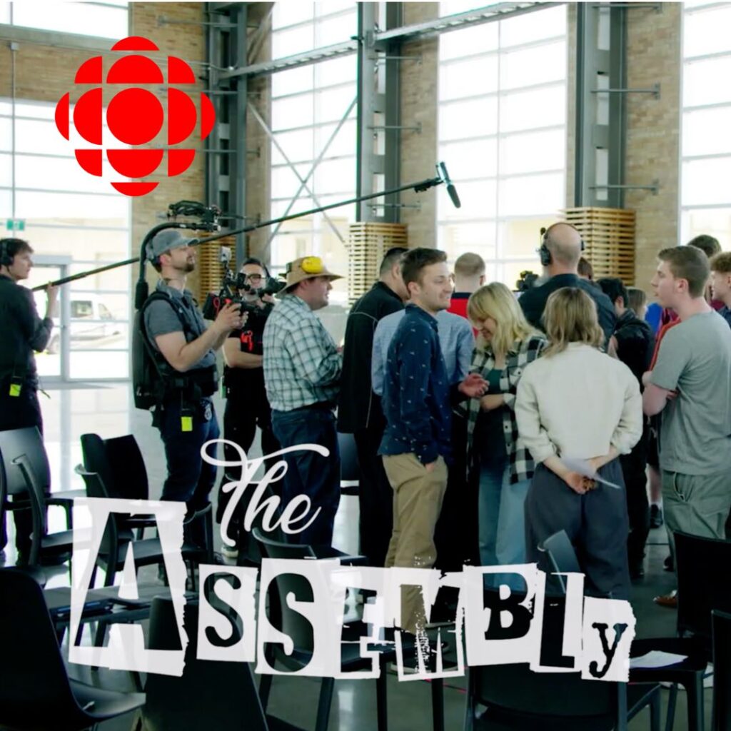 The CBC logo. Title: The Assembly wordmark. A high-ceilinged, bright modern industrial space with a crowd of multi-ethnic, multi-aged people, a boom over their heads and a film crew shooting them.